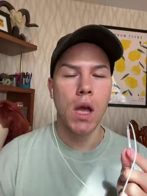 A person with closed eyes, wearing a hat and earphones is holding the earphone cables, against a backdrop of interior items and a lemon art piece.