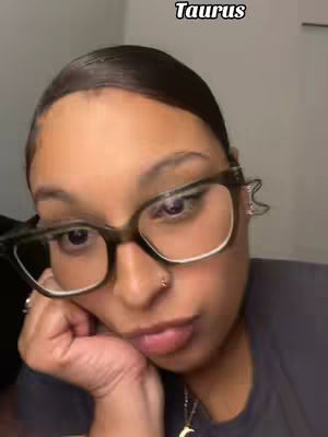 A close-up shot of a woman wearing glasses, with the word "Taurus" in a script font at the top. She is looking down, seemingly resting her head on her hand.