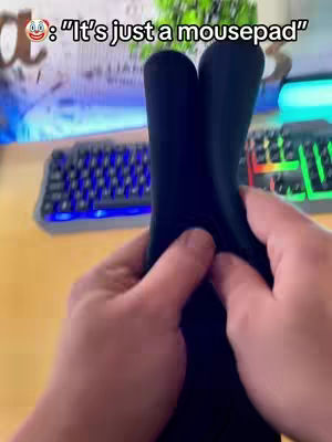 The image shows a first-person perspective of someone holding a black mousepad, the central focus, between their hands. The background is slightly blurred, but includes a keyboard, a monitor, and a plant. The shot is well-lit, with the main focus on the hands and the mousepad.