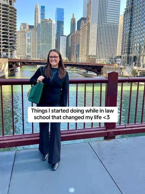 A woman in a dark blazer and pants stands on a bridge, holding a green purse, with a cityscape of tall buildings in the background.