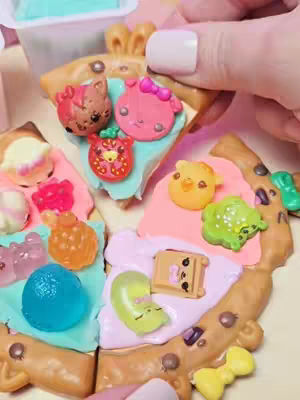 The cover image shows a close-up of a pizza toy, divided into colorful sections with various miniature food and character-shaped toppings. A hand with painted nails is holding a slice of the pizza.