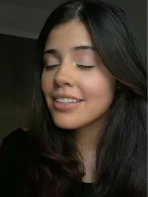 A medium shot of a young woman with dark hair, smiling with her eyes closed, suggesting a moment of happiness or peace. Her skin appears smooth with light makeup. The background is blurred, dark, and out of focus.