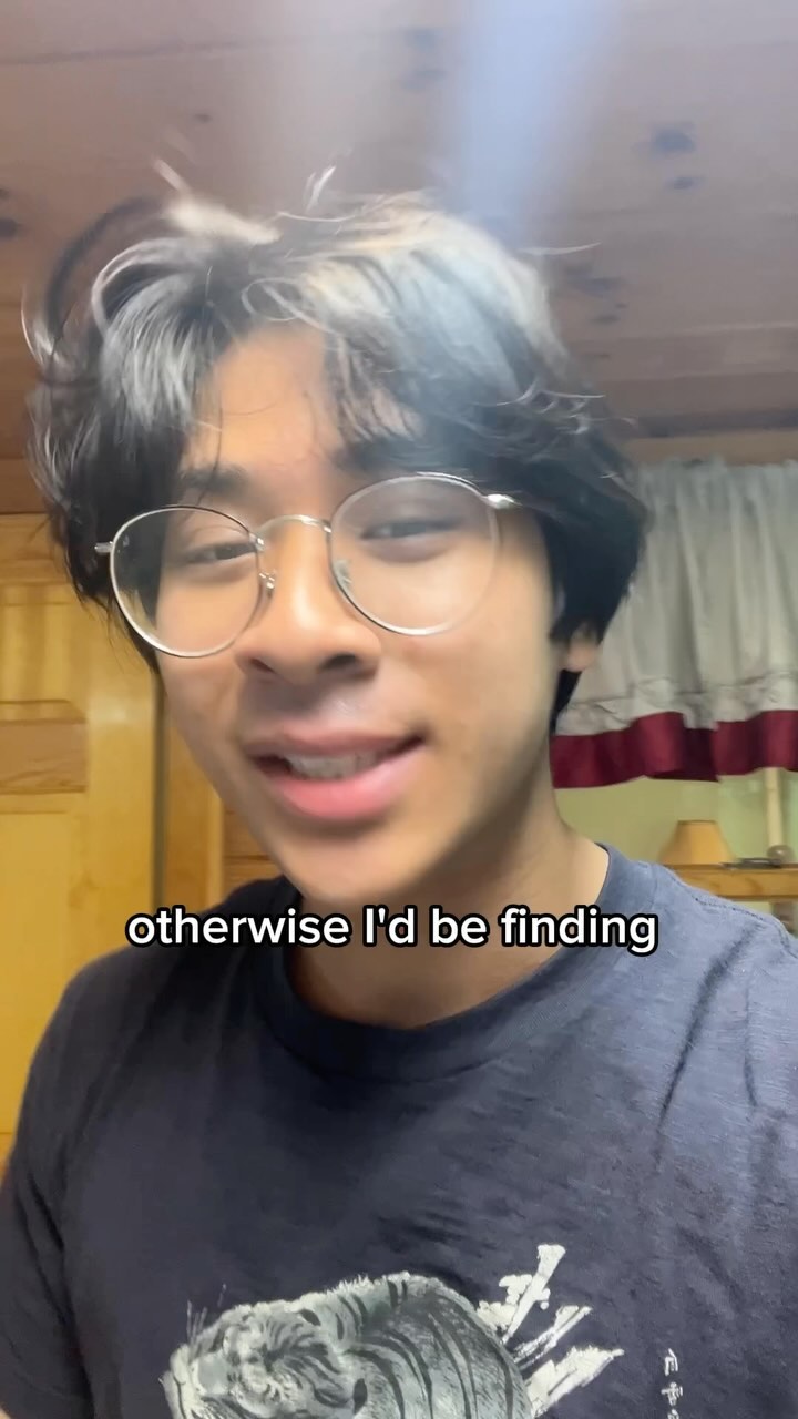 A young man with black hair and glasses is looking at the camera and smiling. The background appears to be a room with a wooden wall and a curtain.