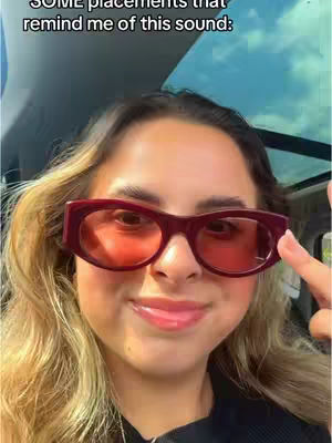 A selfie showing a woman in a car wearing large, red-tinted sunglasses. She is smiling slightly, with her finger up towards the sunglasses on one side. The background is a blue sky visible through the car windows.
