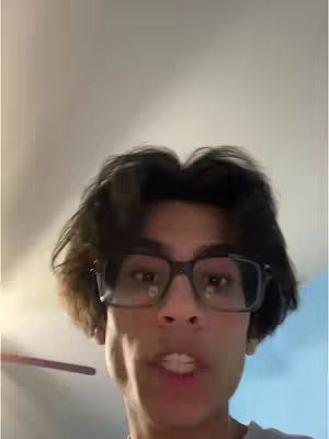 A selfie-style shot featuring a young man in glasses, shot from a low angle. The background is blurred neutral colors, and the man appears to be speaking. The framing is from below.