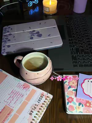 An overhead shot of a desk setup. A laptop and an iPad are visible. On the desk is a cup of coffee, a notebook, a planner, and a lit candle. The color scheme is soft and includes pink, white, and brown.