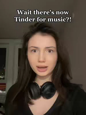 A woman with dark hair and a black shirt is the central figure, with headphones over her neck and a look of surprise. Text overlayed on the image reads, "Wait there's now Tinder for music?!" The background is dark and possibly indoors.