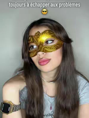 A woman with long brown hair, a gold mask over her eyes, wears a grey t-shirt and gold watch, with a necklace. The background is a plain. The hook text 'Les signes astro qui réussissent toujours à échapper aux problèmes' is at the top.