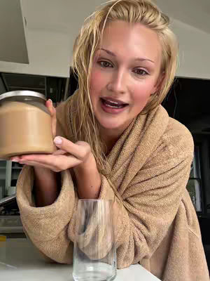A woman with wet blonde hair poses in front of a kitchen counter, holding a jar in one hand and a clear glass on the counter below. She is wearing a beige bathrobe and looking at the camera with a slight smile.