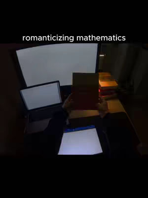 A dimly lit scene featuring a person holding a book, with two monitors and a laptop visible. The text overlay reads 'romanticizing mathematics'. The color palette is muted, with warm lighting illuminating books in the background.