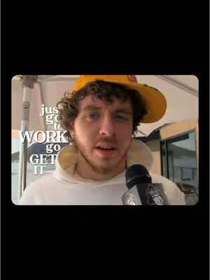A man in a yellow hat and white hoodie speaks into a microphone, with overlay text and a neutral background.