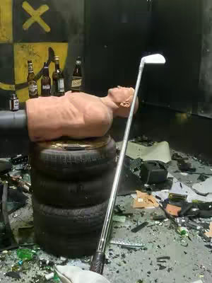 A mannequin torso rests on a stack of tires in a gritty, indoor setting. A golf club leans against the tires, and the foreground is littered with broken glass and debris. Against the black wall, there are beer bottles lined up and some graffiti marks.