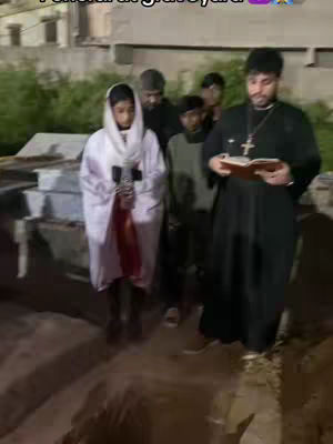 The image depicts a group of people at a graveyard, likely during a funeral ceremony. A person, presumably a priest, in black robes is reading from a book, and another person in a white robe and hood is holding a cross. Several other people stand nearby. The scene is set at dusk or night.