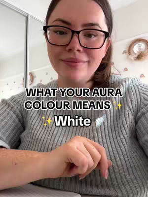 The cover image is a selfie-style shot featuring a woman with glasses. She is wearing a gray sweater. Text overlayed on the image reads "WHAT YOUR AURA COLOUR MEANS" over the word "White". The layout is simple with the woman taking up most of the frame.