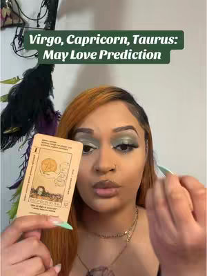 A woman with auburn hair and green eyeshadow holds a tarot card in front of her face. The words "Virgo, Capricorn, Taurus: May Love Prediction" are displayed in a green banner above her head. The background appears to be a neutral wall with some decorative elements.