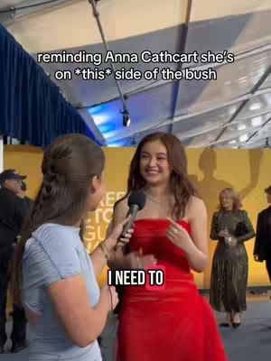 A woman in a blue shirt interviews another woman in a red dress. The background has a yellow horizontal strip with text and blurred figures, and a partial view of a cloudy or tent-like structure overhead.