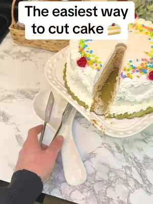 A close-up shot of a cake being sliced with tongs. The cake is white with colorful sprinkles and red rose decorations. The cake sits on a white patterned plate, next to a white handle of a tong. The cake has been cut, with a slice being removed from the cake by the tong. The action occurs over a marble countertop.