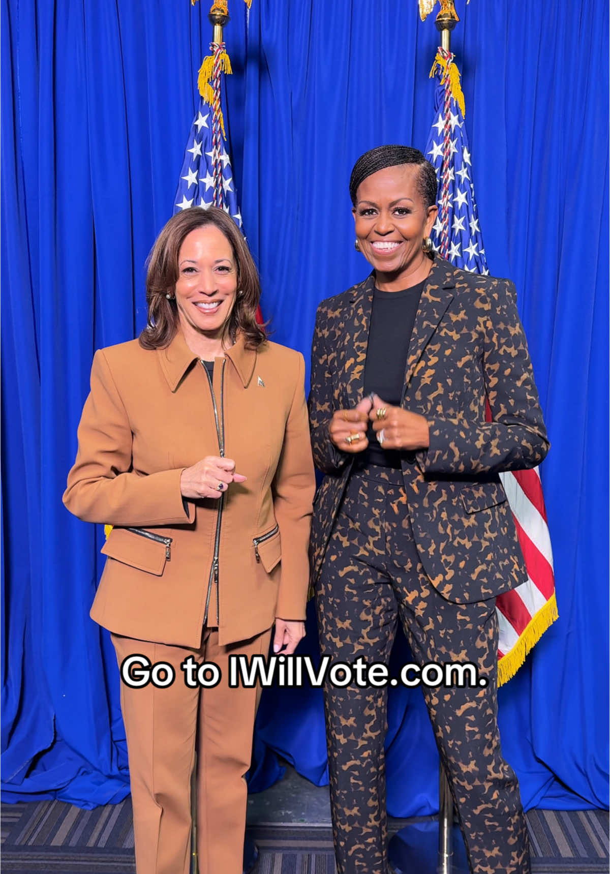 Two women stand side-by-side in front of a blue curtain, with American flags visible between them. Both are smiling and dressed in business attire. Text at the bottom of the image reads 'Go to IWillVote.com.'