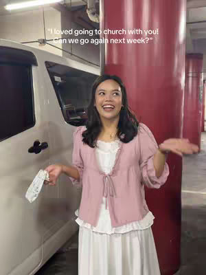 A woman with dark hair smiles broadly, holding a mask and gesturing with her hands in a parking garage setting. She's wearing a pink top over a white dress. Text overlays read a quote about church and the question "Can we go again next week?"
