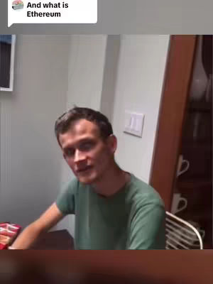 The image features a man with fair skin and short brown hair, wearing a green t-shirt. He is seated indoors, likely in a home, with a neutral gray wall and what looks like a cabinet with open doors behind him. The man is looking towards the camera and speaking. Above, in a speech bubble, is text that reads, "And what is Ethereum."