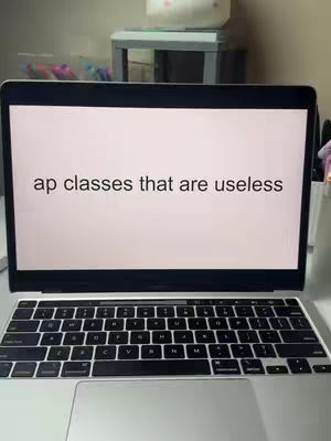 A laptop displaying the text 'ap classes that are useless' on its screen. The laptop is open and positioned on a desk or table, with the keyboard visible. The setting appears to be a well-lit room, possibly a home office or study area. Additional objects are visible in the background, suggesting a personalized workspace.