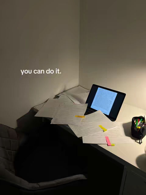 A dimly lit corner showcasing a desk setup for studying or working. A laptop displaying a blue illuminated screen is the central focus, surrounded by scattered papers, some with yellow and pink sticky notes. The room is minimalist with a neutral color palette, emphasizing the workspace ambiance.