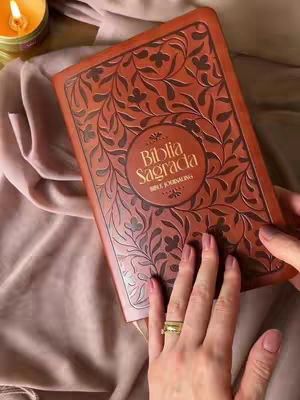 A close-up shot shows a Bible open to a page, held by a person with manicured nails and a ring. The Bible is leather-bound with floral patterns etched into its cover. A candle burns in the upper left corner, casting a warm light over the scene. Soft, draped fabric in shades of pink and beige fills the background, complementing the warm tones of the Bible and candle.
