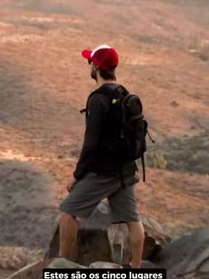 A man stands on a rocky outcrop, facing away from the camera with a red and white hat and backpack. The background is a blurred landscape of golden-brown hills under an overcast sky. Text at the bottom states Este são os cinco lugares (These are the five places)
