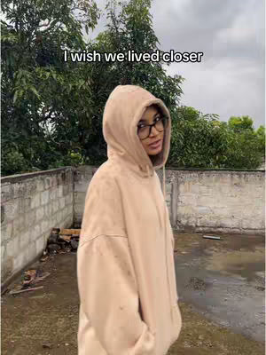 A woman in a beige hoodie and glasses stands outside, tilted slightly, with a background of trees and a cloudy sky. Overlaid text reads 'I wish we lived closer'. The scene appears to be a candid, outdoor shot.
