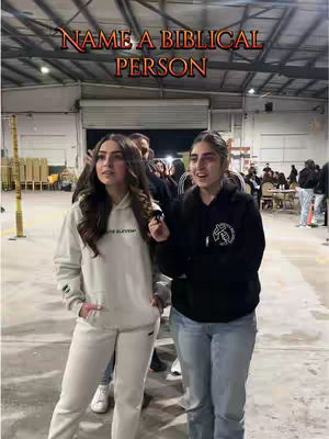 Two young women stand side-by-side in an indoor setting. The woman on the left has brown hair and is wearing a cream-colored hoodie and sweatpants. The woman on the right has dark hair pulled back and is wearing a black hoodie and jeans. Above them, the text "NAME A BIBLICAL PERSON" is displayed in an ornate, orange font.