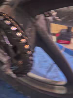 The image is a blurry, dynamic shot focusing on a bicycle chain and wheel. The chain, with its individual links, appears in sharp contrast to the blurred background, potentially indicating movement or speed. The wheel rim is partially visible, and a blue surface (possibly a tarp or fabric) is in the foreground and in the background.