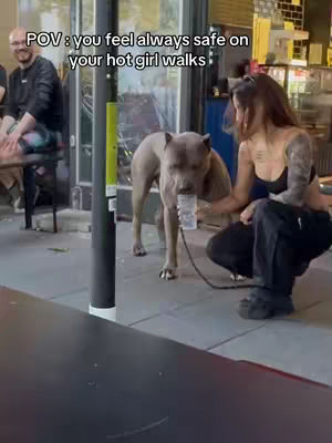 A woman squats, cuddling with a dog that has a clear cup in its mouth. The scene takes place outdoors, with a blurred background featuring parts of a building and another person.