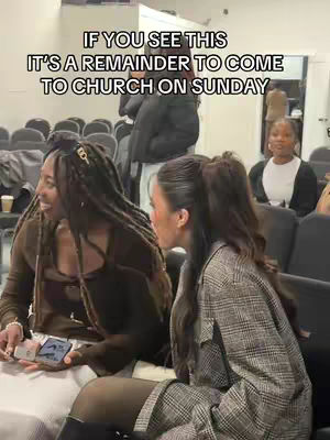 Two women are seated in what appears to be a church setting, with rows of chairs visible in the background. The foreground features a woman with long braids, wearing a brown top, holding a phone with a case. Beside her, another woman, wearing a checkered blazer and with her hair in a ponytail, looks towards her. The text overlay reads, "IF YOU SEE THIS IT'S A REMINDER TO COME TO CHURCH ON SUNDAY."