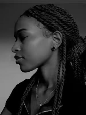 A black and white portrait of a woman in profile. She has her eyes closed and is wearing earrings and a necklace. Her hair is styled in long, dark braids. The background is a simple, dark surface.