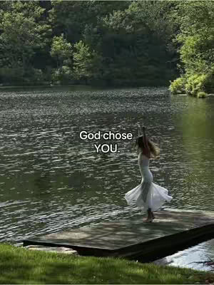 A serene shot featuring a woman in a white dress dancing joyfully on a wooden dock. The background includes a calm lake surrounded by green trees and foliage, illuminated by soft sunlight. The composition is peaceful and natural.