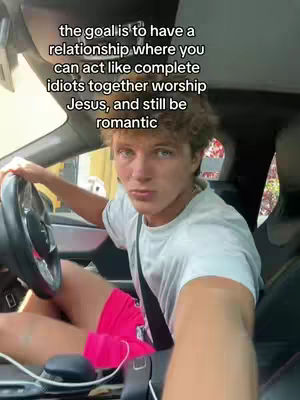 A selfie-style shot of a young man seated in a car. He's holding the steering wheel with one hand, wearing a white t-shirt and pink shorts. Text is overlaid on the image.