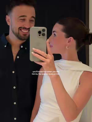 A couple is shown in a selfie, the man on the left looking at the camera and smiling, while the woman on the right holds the phone, gazing at the man. The background is dark.