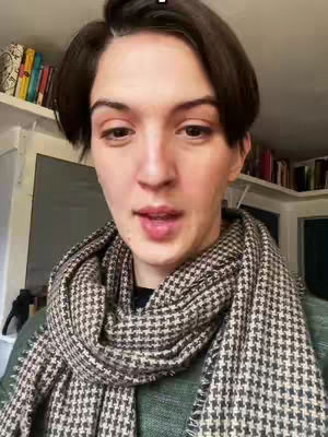 A close-up shot of a woman with short dark hair, wearing a patterned scarf and green top. The background shows bookshelves and the corner of a room. She's looking at the camera and speaking.