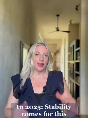 A woman with long, silver hair is centered in the frame, speaking towards the camera. She is in a well-lit hallway or room with warm-toned walls and a dark ceiling fan. Text overlayed at the bottom of the image announces, "In 2025: Stability comes for this."