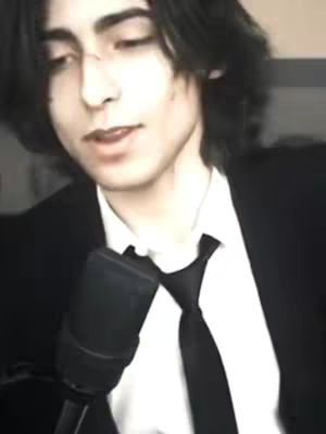 A medium shot of a young man with dark hair, wearing a black suit jacket and a white shirt with a black tie, holding a black microphone. The background is out of focus.