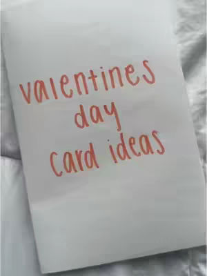 A white sheet of paper with the words 'valentines day card ideas' written in red marker. The paper is slightly wrinkled, and the background is a soft, blurred white and gray.