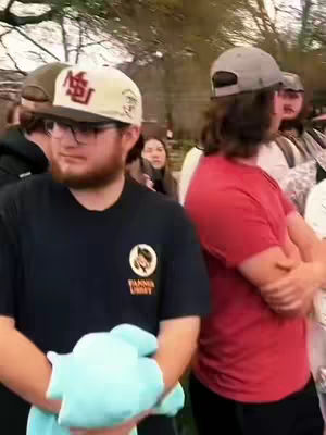 The cover image shows a group of people standing outside, with two men prominently in the foreground. The man on the left is wearing a dark blue shirt, glasses, and a baseball cap with text on it. He holds a light blue object in his hands. The second man on the right is wearing a red shirt and a gray cap. The background has a blur of trees and a few other people.