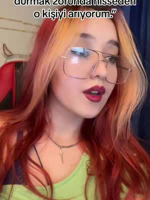 A medium shot of a young woman with fiery red and blonde hair, wearing large-framed glasses and a light green top, looking towards the camera with a slight smile. She has red lipstick and a silver necklace with a cross pendant. The background is blurred, but hints at a room setting.
