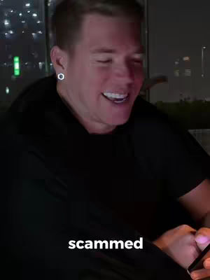 A man in a black hoodie is smiling widely, with a window and city lights visible in the background.