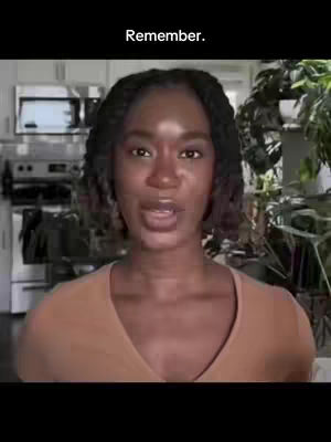 A woman with dark hair and brown skin is the central focus, set against a blurred domestic backdrop. The woman is positioned in the foreground, facing the camera directly and speaking. Her expression appears engaged. The background is a kitchen space, with visible elements including cabinetry, a stovetop, and various potted plants, suggesting a well-lit, indoor environment. A text overlay at the top reads 'Remember'. The overall composition is straightforward, emphasizing the woman's communication.