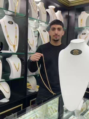 A man in a black sweater holds a gold necklace in a jewelry store, with numerous other gold necklaces and jewelry displays in the background. The setting seems to be a shop, with glass shelves and various gold items on display.