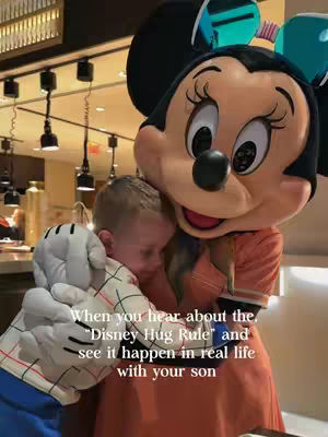 A person is hugging a Minnie Mouse character at what appears to be a crowded restaurant or hotel. The Minnie Mouse character is prominently featured, with a large head, black ears, and a red dress. The person is a young child, with their face partially obscured against Minnie's costume with the overlayed text.