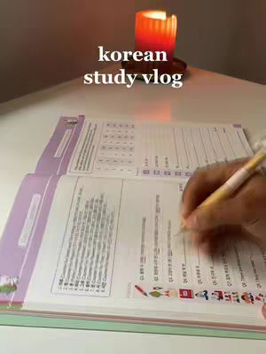 The cover image shows an open Korean language study book on a white desk. A hand is seen writing with a yellow pen on the page. Above the book, there's a lit orange candle. The words 'korean study vlog' are overlaid in white text.