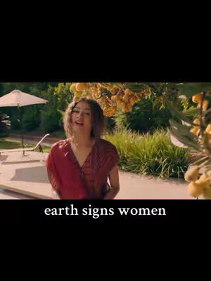 A woman stands outside near a pool. She has her head turned upwards, and she is wearing a red top. The background is blurred, but it appears to be a mix of green foliage and buildings. Text overlayed at the bottom of the image says "earth signs women".