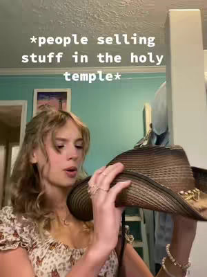 A young woman with light skin is shown in a selfie-style shot, looking slightly to her left. She holds a brown hat in front of her, with a light-colored top, and a few rings visible on her fingers. The background is a mix of light blue and white, suggesting an interior setting.